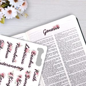 May include: A sheet of adhesive Bible index tabs featuring black cursive text on a white background with pink floral accents. The tabs include labels for Genesis, Exodus, Leviticus, Numbers, Deuteronomy, Joshua, Ruth, and 1 Samuel, placed on a Bible.
