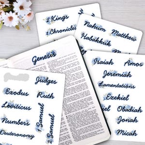 May include: A collection of white sticker sheets featuring the names of various books of the Bible printed in a dark blue cursive font, each accented with small blue floral illustrations, displayed alongside an open book with printed text.