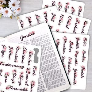 Könnte beinhalten: Eine Sammlung weißer, selbstklebender Bibelregister mit schwarzer Schreibschrift und rosa Blumenmotiven. Die Register sind auf Bögen angeordnet und liegen neben einer aufgeschlagenen Bibel, auf deren Seite Genesis steht.