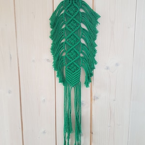 Peut inclure: Une suspension murale en macramé verte avec un anneau en bois en haut. La suspension a un motif en losange et de longues franges.