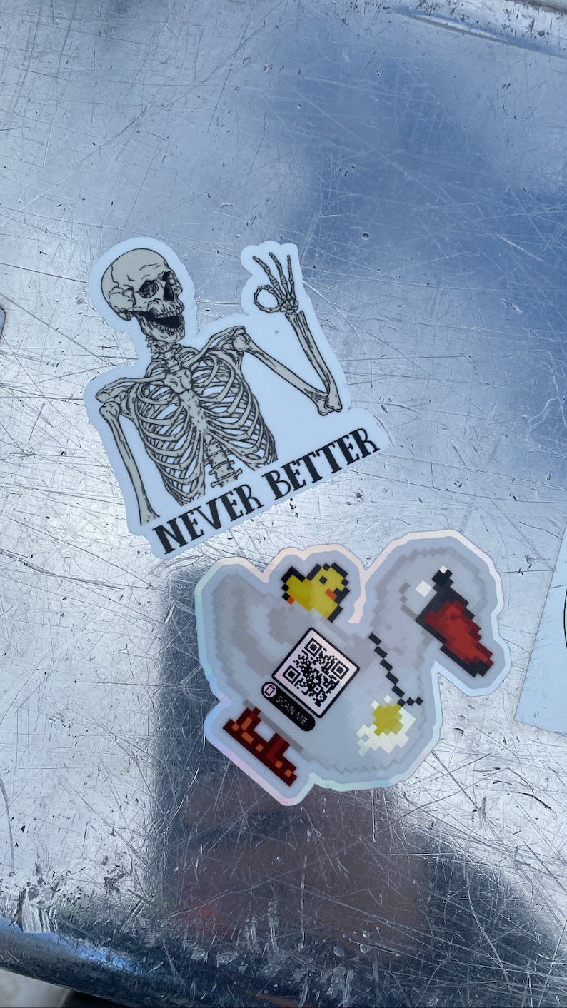 May include: Two stickers on a silver surface. The first sticker is a skeleton with the text "NEVER BETTER". The second sticker is a pixelated image of a duck with a QR code and the text "SCAN ME".