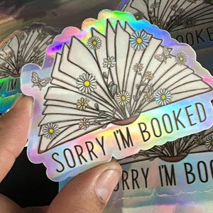 May include: A holographic sticker featuring a book design with the text "Sorry I'm Booked". The book is surrounded by flowers and a butterfly.