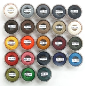 Peut inclure: Une collection de 24 bobines de fil de différentes couleurs, notamment blanc, gris, marron, orange, rouge, bleu, vert et jaune. Chaque bobine est étiquetée avec un numéro, comme "V01-100", "V01-115" et "V01-155".