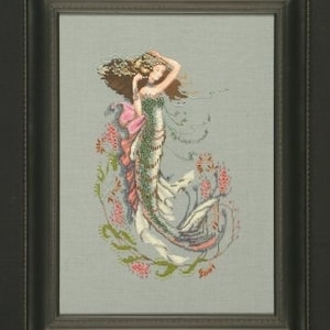 Puede incluir: Un bordado de punto de cruz de una sirena con el pelo largo y suelto y una cola verde. Está rodeada de flores rosas y verdes. El bordado está enmarcado en un marco marrón oscuro.