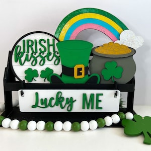 May include: A St. Patrick's Day themed wooden display with a black frame. It features a rainbow, pot of gold, leprechaun hat, and signs that read "Irish Kisses" and "Lucky Me". A string of green and white beads is at the base.