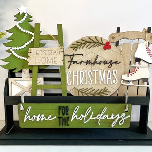 May include: A farmhouse Christmas themed tiered tray with a green Christmas tree, a ladder with the text "Let's Stay Home", a round sign that says "Farmhouse Christmas", a wooden sled with ice skates, and a sign that says "Home for the Holidays".