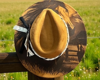 May include: A brown cowboy hat with a horse graphic, a tan crown, and a white lace trim. The hat is decorated with a black ribbon and white lace. The hat is hanging on a wooden fence post.