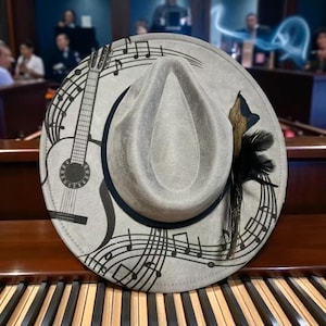 May include: Gray fedora hat with a black brim, decorated with a black guitar outline, musical notes, and a feather accent. The hat is displayed on a piano keyboard.