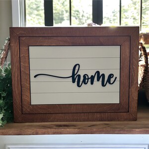 May include: A wooden framed sign with a white background and black script lettering that reads "home".