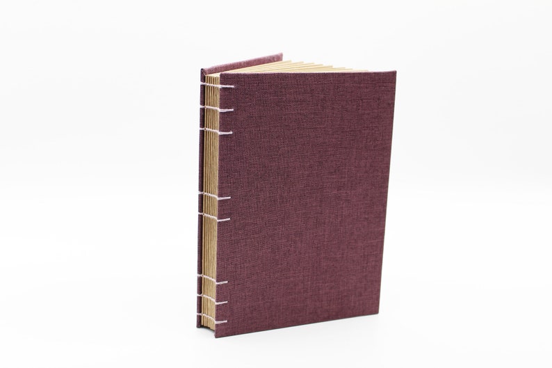 May include: A burgundy linen-covered journal with a spine sewn with white thread. The journal is closed and standing upright on a white surface.