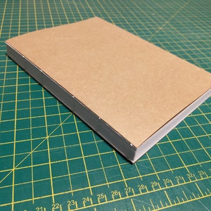 Puede incluir: Un libro cubierto de cartón marrón con lomo gris y páginas blancas. El libro está sobre una estera de corte verde.