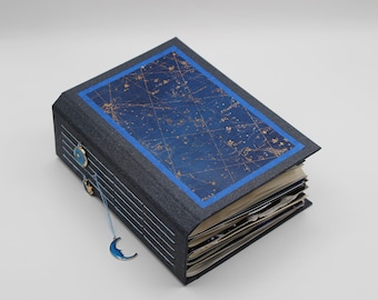 Op de afbeelding: Een donkerblauw dagboek met een sterrenkaart op de omslag. De omslag heeft een blauwe rand en gouden accenten. Het dagboek heeft een blauwe halve maan-hanger.
