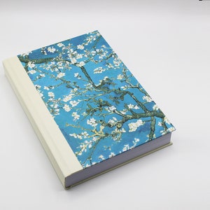 May include: A blue hardcover journal with a white border and a floral design on the cover. The design features white flowers and green branches on a blue background.
