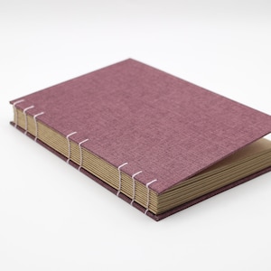 Puede incluir: Un libro cerrado con una cubierta de lino malva y un lomo con costuras visibles. El libro está sobre una superficie blanca.