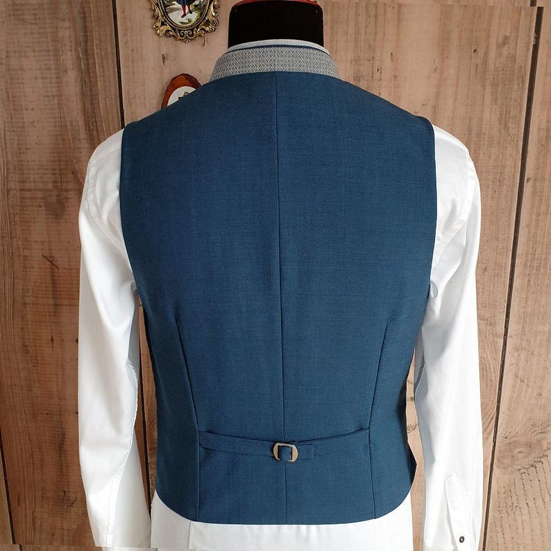 Puede incluir: Un chaleco azul turquesa con una hebilla dorada en la espalda. Se lleva sobre una camisa blanca de manga larga. El chaleco tiene un corte entallado y un dise&ntilde;o cl&aacute;sico.