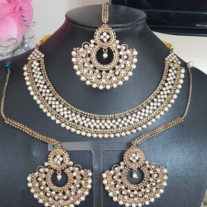 May include: A gold-tone bridal jewelry set with white pearls and clear rhinestones. The set includes a necklace, earrings, and a maang tikka.