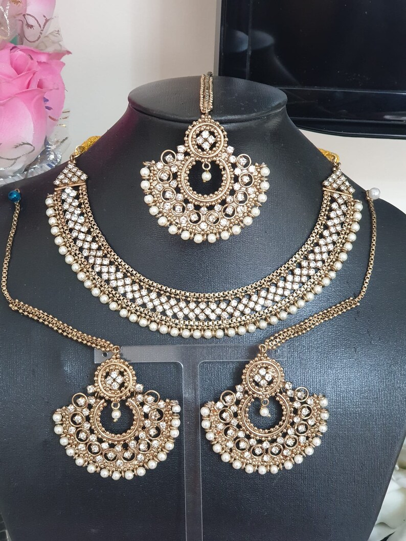 May include: A gold-tone bridal jewelry set with white pearls and clear rhinestones. The set includes a necklace, earrings, and a maang tikka.