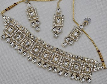 May include: A gold necklace and earrings set with clear cubic zirconia stones. The necklace features a row of square-shaped stones with teardrop-shaped stones hanging below. The earrings are similar in design, with a square-shaped stone and teardrop-shaped stones hanging below.