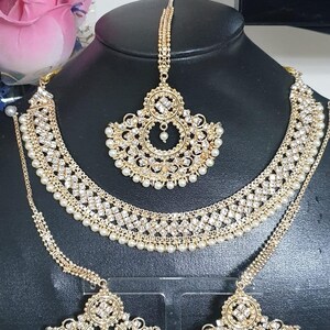May include: A gold-tone bridal jewelry set with white pearl accents. The set includes a necklace, earrings, and a maang tikka.