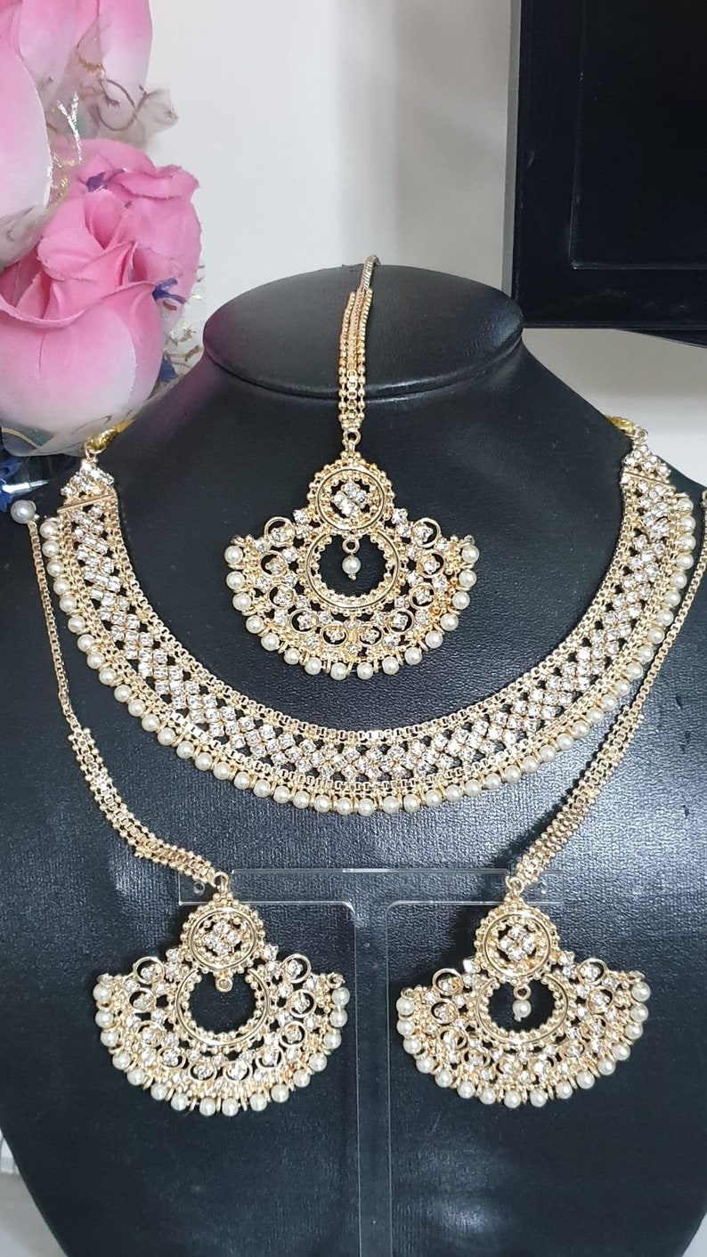 May include: A gold-tone bridal jewelry set with white pearl accents. The set includes a necklace, earrings, and a maang tikka.