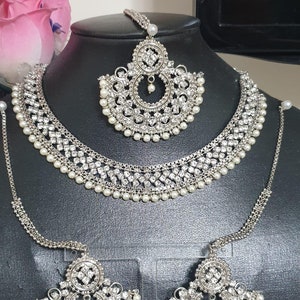 May include: A silver and white bridal jewelry set with a necklace, earrings, and a maang tikka. The set is adorned with sparkling rhinestones and pearls.