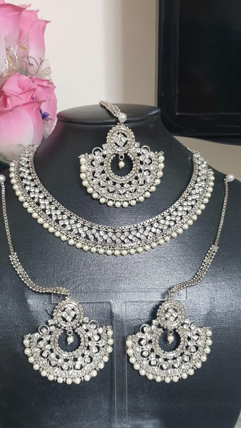 May include: A silver and white bridal jewelry set with a necklace, earrings, and a maang tikka. The set is adorned with sparkling rhinestones and pearls.