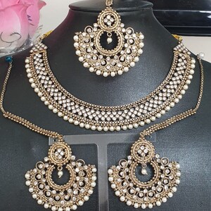 May include: A gold-tone Indian bridal jewelry set with white pearl accents. The set includes a necklace, earrings, and a maang tikka.