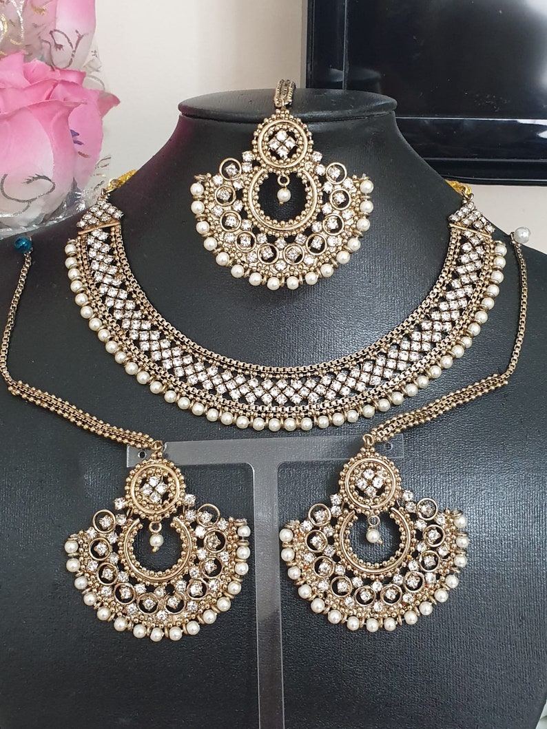 May include: A gold-tone Indian bridal jewelry set with white pearl accents. The set includes a necklace, earrings, and a maang tikka.