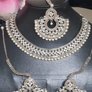May include: A silver bridal jewelry set with white pearls and clear rhinestones. The set includes a necklace, a maang tikka, and a pair of earrings.