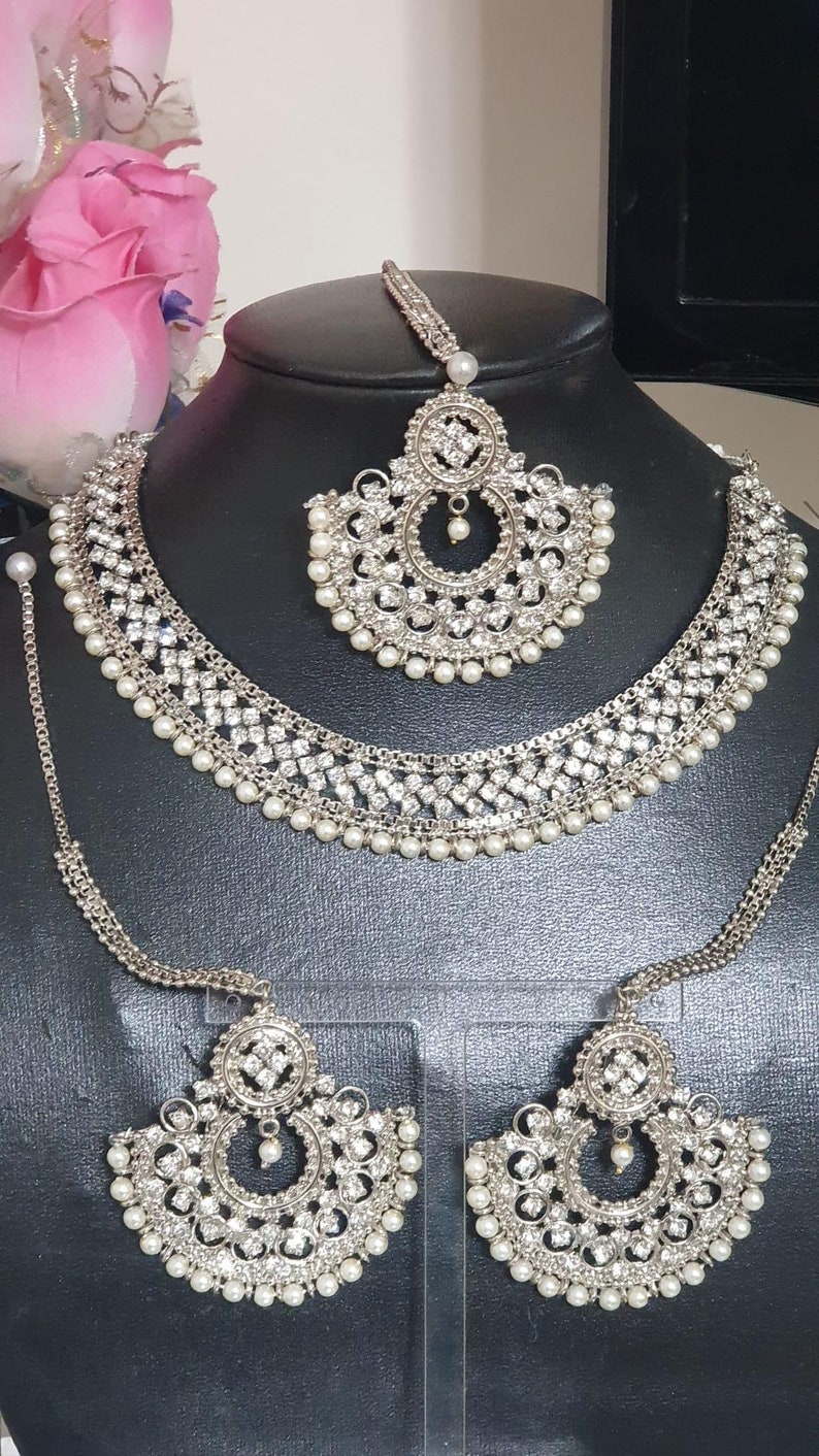 May include: A silver bridal jewelry set with white pearls and clear rhinestones. The set includes a necklace, a maang tikka, and a pair of earrings.