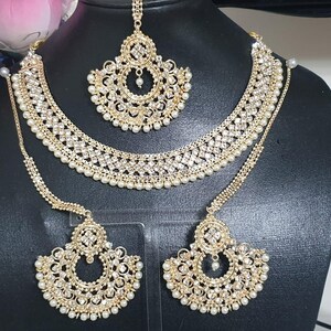 May include: A gold-tone bridal jewelry set with white pearl accents. The set includes a necklace, earrings, and a maang tikka.