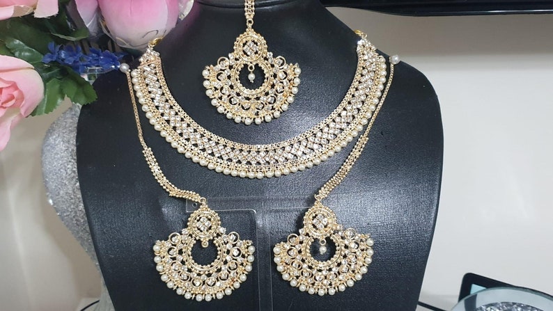 May include: A gold-tone bridal jewelry set with white pearl accents. The set includes a necklace, earrings, and a maang tikka.