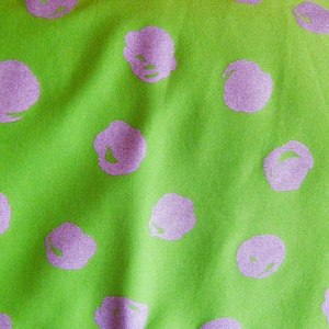 以下が含まれることがあります： ライムグリーンの生地のクローズアップ。大きなラベンダー色の花の形が散りばめられています。生地は柔らかい質感で、ほのかな光沢があり、視覚的に魅力的なパターンを作り出しています。