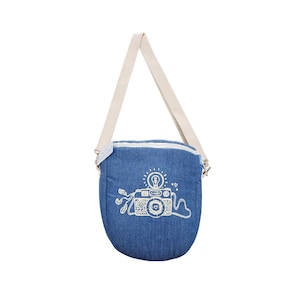 Puede incluir: Bolso bandolera de mezclilla azul con correa blanca. El bolso presenta un diseño de cámara bordada en blanco con la palabra "smile" y elementos decorativos. El bolso tiene cierre de cremallera y herrajes plateados.