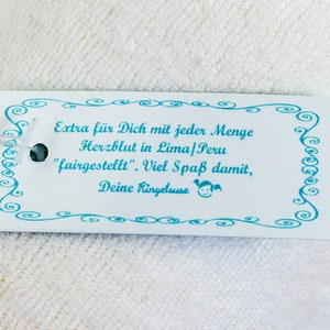 May include: A rectangular white paper tag with a light blue decorative swirl border and a light blue ribbon. The tag features the text: Extra f&uuml;r Dich mit jeder Menge Herzblut in Lima/Peru "fairgestellt". Viel Spa&szlig; damit, Deine Ringelouse.