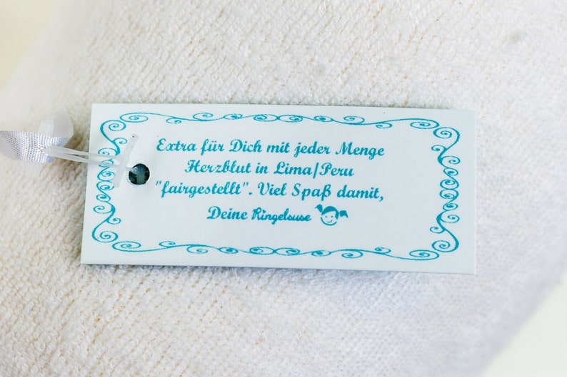 May include: A rectangular white paper tag with a light blue decorative swirl border and a light blue ribbon. The tag features the text: Extra f&uuml;r Dich mit jeder Menge Herzblut in Lima/Peru "fairgestellt". Viel Spa&szlig; damit, Deine Ringelouse.