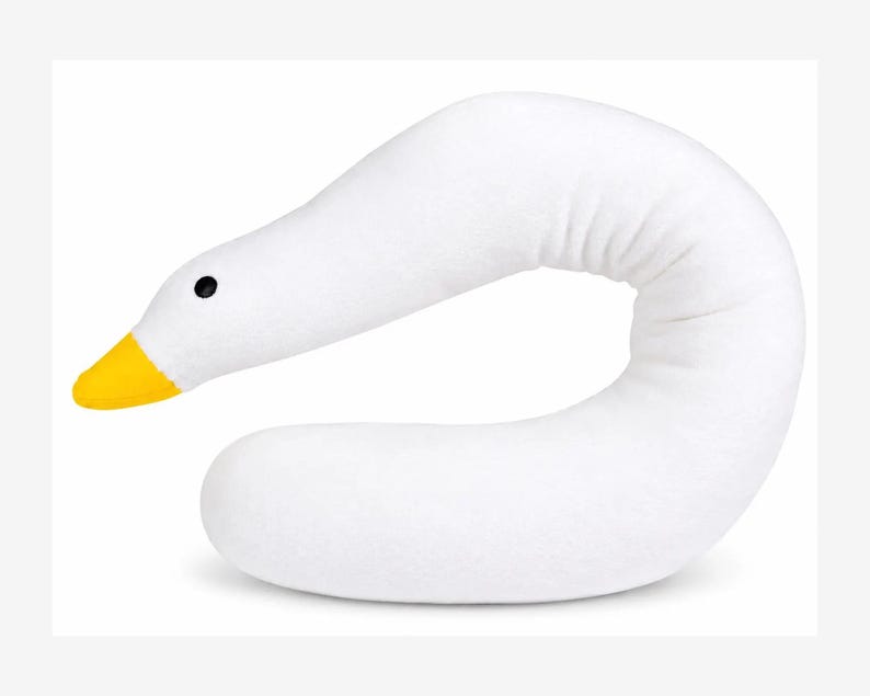May include: A white plush pillow shaped like a long, curved goose neck. The pillow features a bright yellow beak and a small, round black eye. The soft, elongated body curves into a U-shape, mimicking the graceful posture of a goose's neck and head.