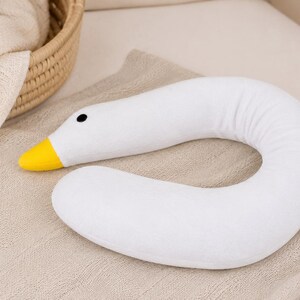 May include: A white, U-shaped neck pillow designed to resemble a goose. The pillow features a soft, plush texture, a bright yellow beak, and a small, round black eye. The curved body of the pillow is smooth and uniform in its white color.
