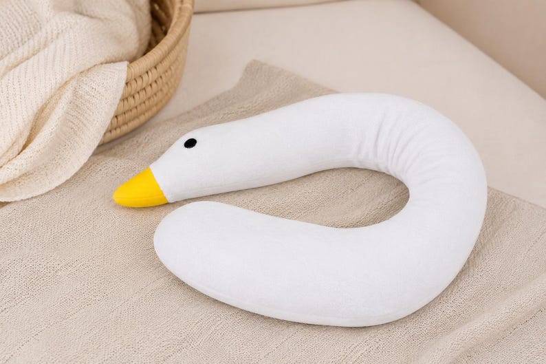 May include: A white, U-shaped neck pillow designed to resemble a goose. The pillow features a soft, plush texture, a bright yellow beak, and a small, round black eye. The curved body of the pillow is smooth and uniform in its white color.