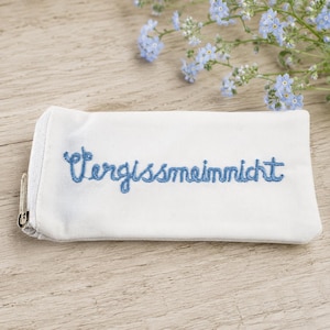Könnte beinhalten: Ein weißes, rechteckiges Stoffetui mit der blauen Stickerei Vergissmeinnicht. Das Etui verfügt über einen silberfarbenen Metallreißverschluss an der linken Seite. Das Design wird durch kleine hellblaue Blüten oberhalb des Etuis ergänzt.