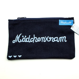Peut inclure: Pochette zippée bleu marine avec le mot "Mädchenkram" brodé en script bleu clair. La pochette a une petite étiquette bleue avec du texte et trois petits cœurs bleu clair.