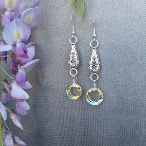 May include: Silver dangle earrings with a teardrop-shaped charm and a round crystal charm. The crystal charm is a light yellow color with a blue iridescent sheen.