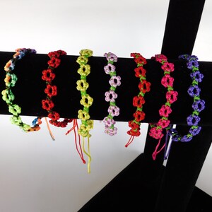 May include: Assortment of colorful friendship bracelets with flower designs. The bracelets are made with colorful embroidery floss and are displayed on a black stand.