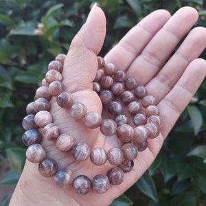 May include: A brown and gold sunstone beaded bracelet with a shimmery finish. The bracelet is made of round beads and is shown on a hand.