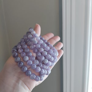May include: A stack of lavender amethyst gemstone beads, each bead is round and smooth. The beads are arranged in a circular pattern, creating a layered effect.