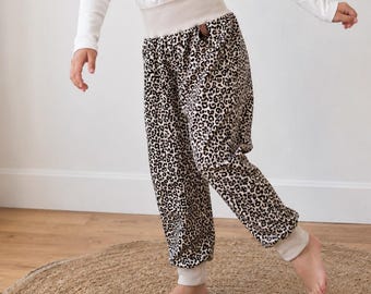 Könnte beinhalten: Ein junges Mädchen trägt ein weißes Langarmshirt mit Rüschendetails und eine Hose mit Leopardenmuster. Die Hose hat einen beigen Bund und Bündchen. Sie steht auf einem runden, gewebten Teppich.
