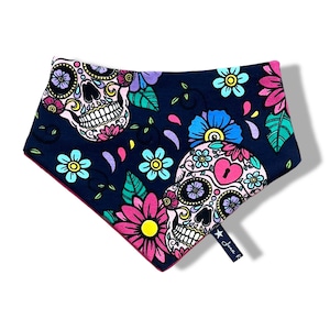 Könnte beinhalten: Ein schwarzes Bandana mit einem rosa, blauen und gelben Blumenmuster und zwei Totenkopf-Designs. Das Bandana hat einen rosa Rand und ein kleines sternförmiges Etikett mit dem Text "Luna K" darauf.