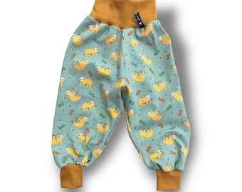 Op de afbeelding: Babybroek met een blauwgroene achtergrond en een patroon van gele luiaards en groene bladeren. De manchetten en tailleband zijn mosterdgeel. De broek heeft een hoge taille en een relaxte pasvorm.