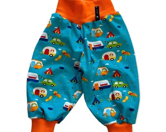 Op de afbeelding: Babybroek met een turquoise achtergrond met een campingthema met campers, tenten en auto's. De broek heeft een oranje tailleband en manchetten. Een klein zwart label met witte tekst is zichtbaar op de tailleband.