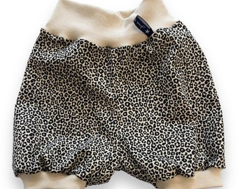 Könnte beinhalten: Beige Shorts mit schwarzem Leopardenmuster, weißem elastischem Bund und Bündchen. Die Shorts haben ein kleines schwarzes Etikett mit einem weißen Stern und dem Text "Junior" darauf.